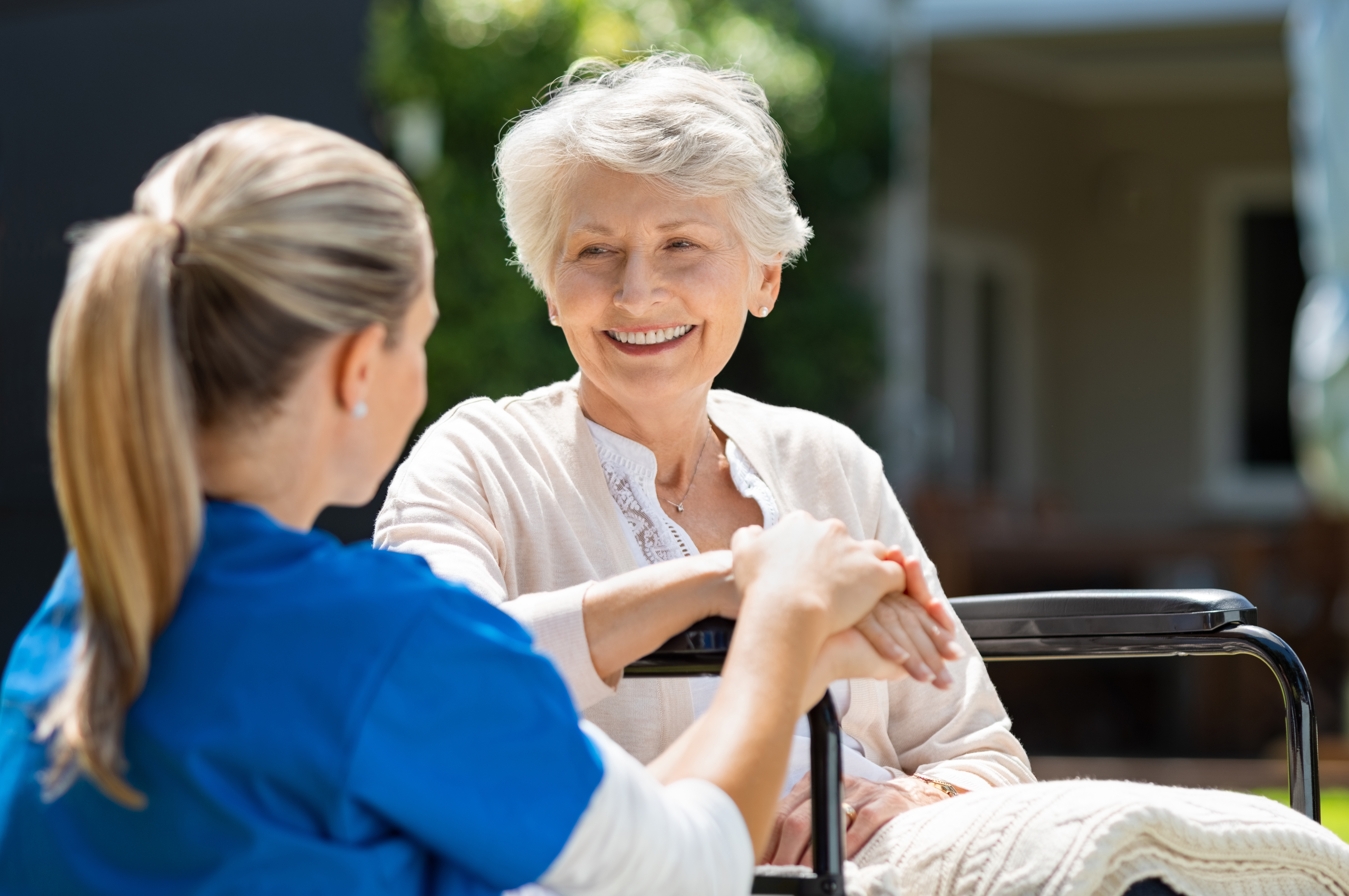 Smiling senior patient sitting on wheelchair with nurse supporting her. Doctor looking at elderly patient on a wheelchair in the garden. Nurse holding hand of mature woman outside pension home.