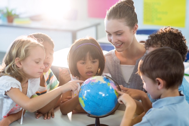 Smiling female teacher looking at schoolchildren touching globe in classroom
